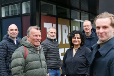 Kuusi hymyilevää henkilöä seisoo ulkona modernin rakennuksen edustalla. Henkilöt ovat pukeutuneet lämpimiin vaatteisiin, mikä viittaa viileään säähän. Heidän takanaan oleva rakennus ja ilmeet viestivät yhteistyöstä, hyvästä tunnelmasta ja todennäköisesti työ- tai projektitapaamiseen liittyvästä hetkestä.