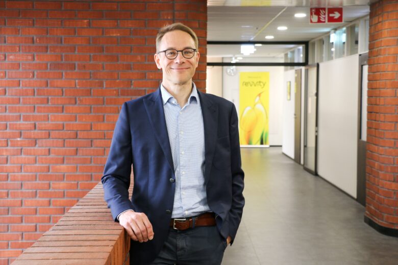 Janne Seppälä, Director of Product Development for the Reproductive Health unit, stands indoors next to a brick wall, smiling at the camera. He is wearing a navy blue blazer, a light blue dress shirt, and dark trousers. In the background, a modern office corridor and a yellow Revvity poster are visible.