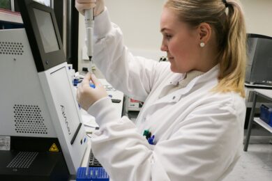 Laboratoriotyöntekijä valkoisessa takissa pipetoi näytettä koeputkeen modernissa laboratoriossa. Hänellä on vaaleat hiukset poninhännällä, helmikorvakorut ja suojakäsineet. Työpisteessä on Revvity-laboratoriolaite ja sininen pipettiteline.