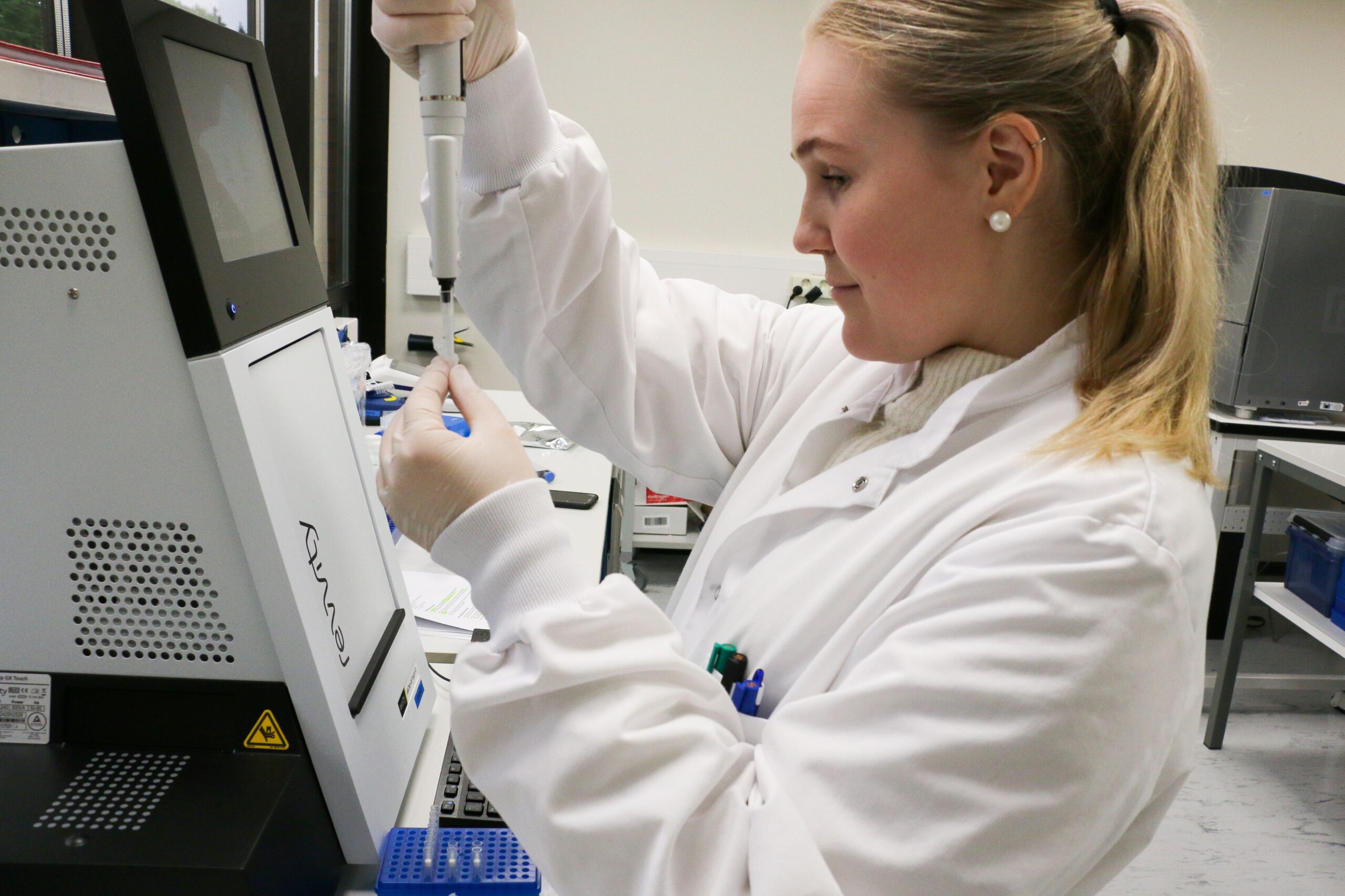 Laboratoriotyöntekijä valkoisessa takissa pipetoi näytettä koeputkeen modernissa laboratoriossa. Hänellä on vaaleat hiukset poninhännällä, helmikorvakorut ja suojakäsineet. Työpisteessä on Revvity-laboratoriolaite ja sininen pipettiteline.
