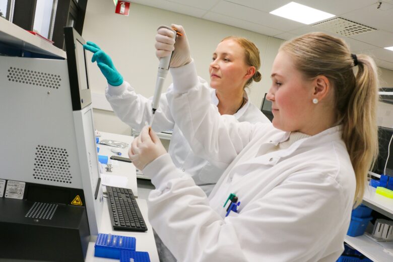 Kaksi laboratoriotyöntekijää valkoisissa takissa työskentelee pipetin ja laboratoriolaitteen kanssa. Etualan henkilö pitää koeputkea ja pipetoi näytettä, kun taas taustalla oleva kollega seuraa prosessia ja käyttää laitteen kosketusnäyttöä. Molemmilla on suojakäsineet, ja työympäristö on moderni ja siisti.