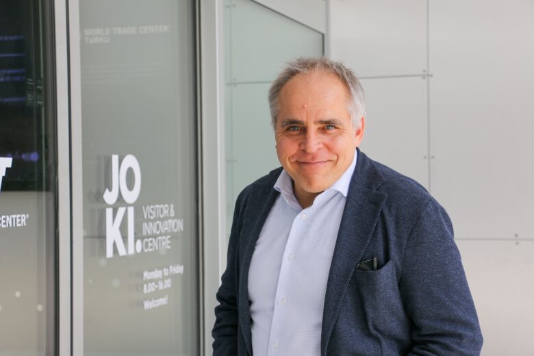 Hymyilevä mies seisoo Joki Visitor & Innovation Centren sisäänkäynnin edustalla Turussa. Hänellä on päällään sininen bleiseri ja valkoinen kauluspaita.