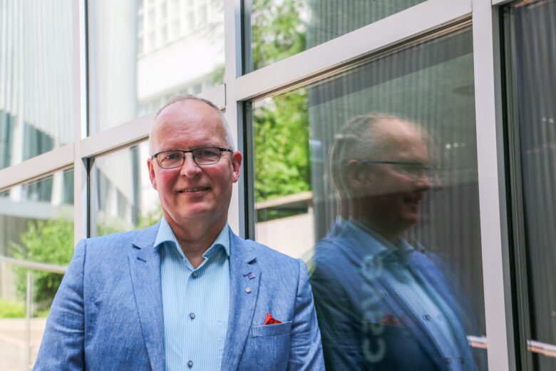 Työ- ja elinkeinoministeriön (TEM) Harri Länsipuro seisoo lasiseinän edessä hymyillen. Hänellä on vaaleansininen puku ja silmälasit, ja hänen heijastuksensa näkyy ikkunalasista