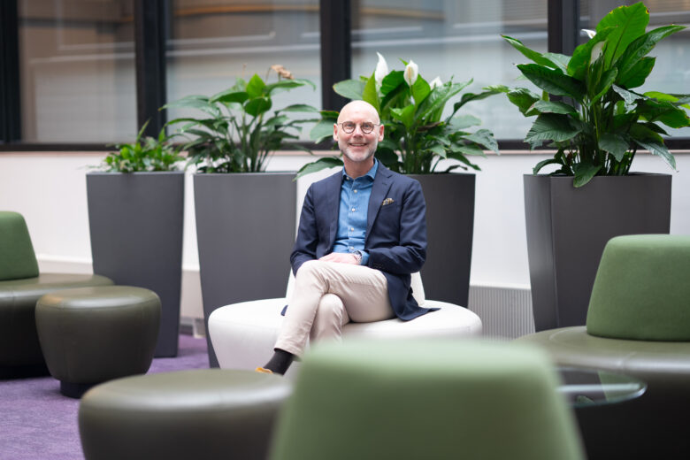 Olli Kuussaari, CEO of Vetcare, sits cross-legged on a white couch in a modern office lounge. He is smiling and looking at the camera. He is wearing a navy blue blazer, denim shirt, and beige trousers. Behind him are large green plants and windows. In the foreground are green and dark green lounge chairs and a purple carpet.