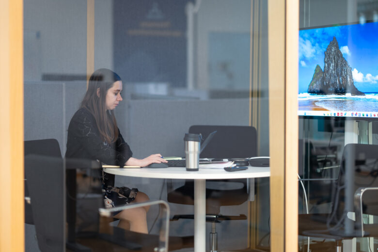 A woman working on a laptop in a small meeting room. She is seated at a round table, focused on her screen. On the table are a notebook, a thermos mug, and a large display screen showing a beach-themed wallpaper. The photo is taken through a glass wall.