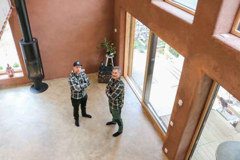 Pure Home Nordic founders Niklas Sampalla and Jari Petäjämäki stand in the living room of a straw house with large windows and clay-plastered walls.