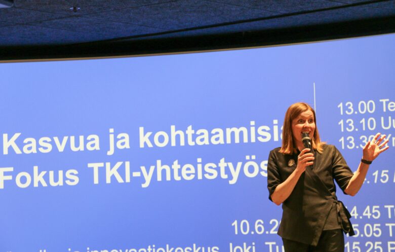 A speaker from Business Turku addresses the audience at the “Growth and Encounters – Focus on RDI Collaboration” seminar, held at the Visitor and Innovation Centre Joki in Turku in June 2025. A large blue screen behind her displays the event title and program details.