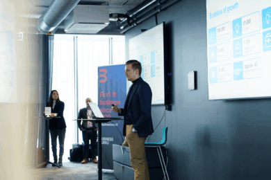 Mies pitää puhetta, takana kaksi näyttöä.