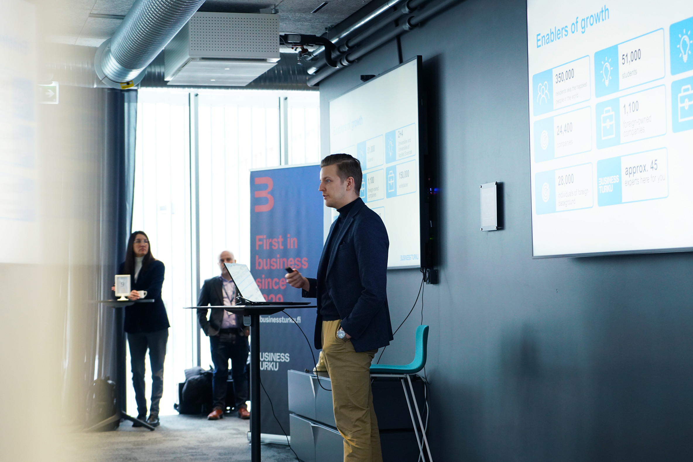 Mies pitää puhetta, takana kaksi näyttöä.
