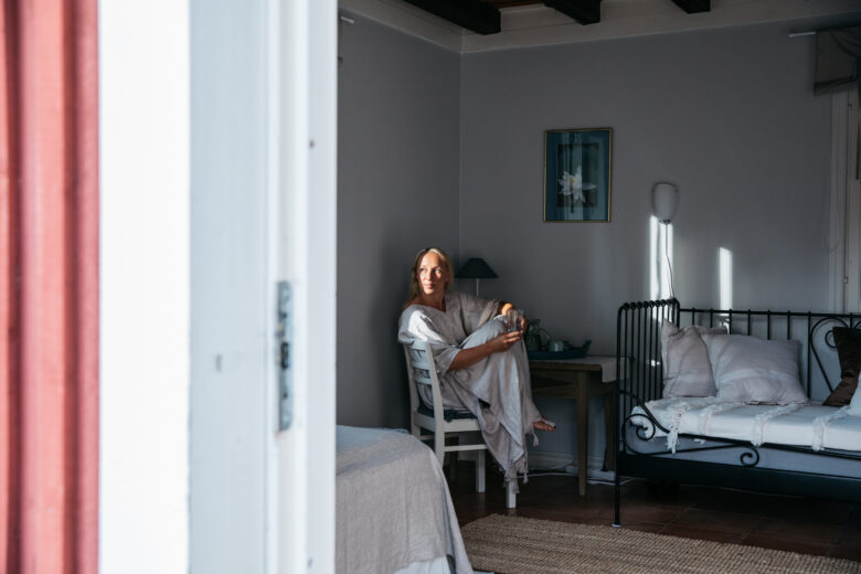 Hanna Kallioniemi istuu sisällä valoisassa huoneessa nojatuolissa ja lukee, ympärillä pehmeäsävyinen sisustus ja päivävalo.”