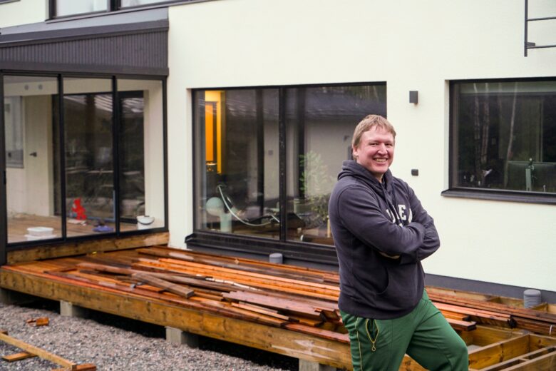 Mikko Hällfors seisoo nykyaikaisen omakotitalon terassin työmaan edessä, taustalla isot ikkunat ja rakennusmateriaaleja.