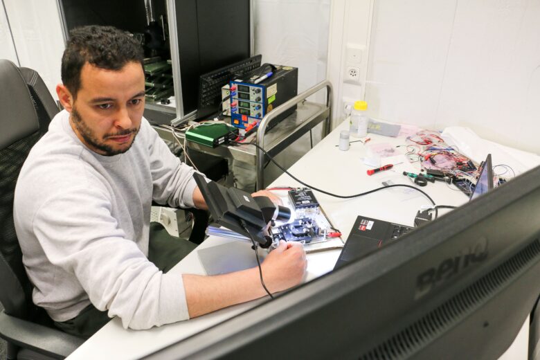 Työpisteessä henkilö työskentelee elektroniikkakomponenttien parissa käyttäen mittauslaitteita ja työvaloa.