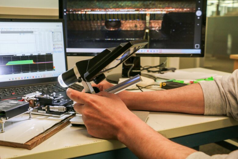 Työpöydällä oleva laite tutkitaan käsikäyttöisellä optisella mittalaitteella, ja taustalla näkyy analyysiohjelmia tietokoneen näytöillä.