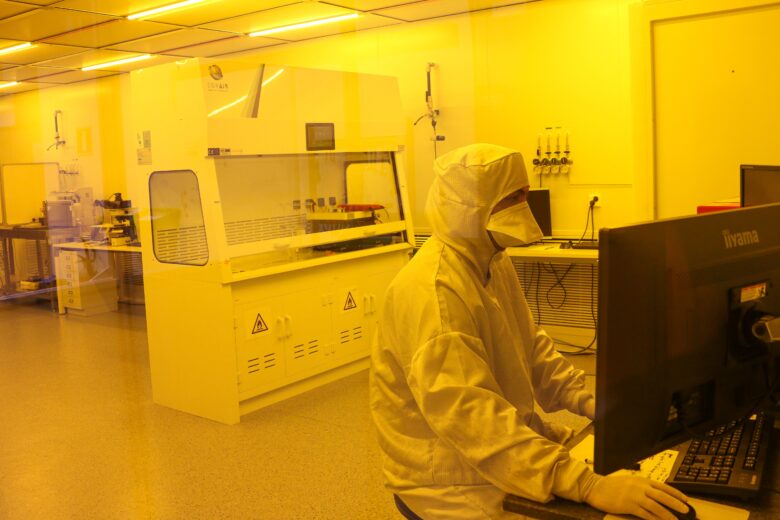 In a yellow-lit cleanroom, a person wearing protective clothing works at a computer next to industrial equipment.