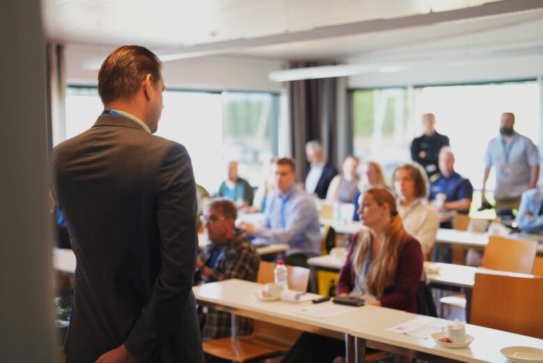 Kuvan etualalla selin kameraan Mikko Pänkäälä puhumassa pienelle yleisölle, jotka istuvat pitkien pöytien äärellä.
