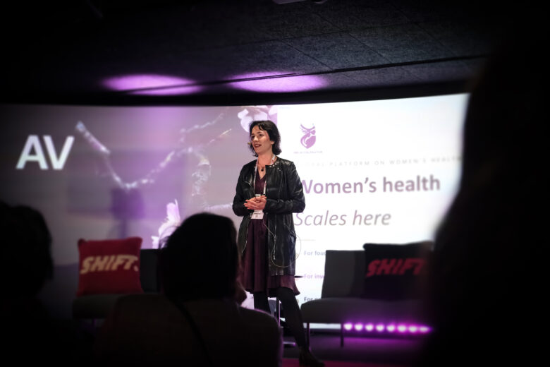 Anastasiya Markvarde stands on stage at the SHIFT event, presenting a slide about women’s health spending to an audience.