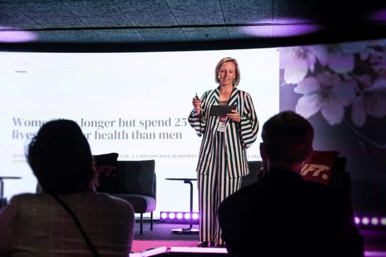 Aura Pyykönen presents on stage at the SHIFT event, with a slide reading “Women’s health scales here” in the background.