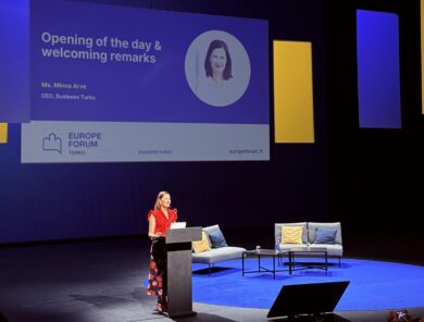 Business Turun toimitusjohtaja Minna Arve pitää avauspuheenvuoroa Eurooppa-foorumin lavalla 2025 Turun kaupunginteatterissa. Taustalla suurella näytöllä näkyvät avauksen otsikko "Opening of the day & welcoming remarks", Minna Arven nimi sekä Europe Forum Turku -tapahtuman tunnukset. Lavalla on puhujakorokkeen lisäksi paneelikeskustelua varten sohvia, pöytiä ja istuinryhmä sinisellä valaistulla näyttämöllä.