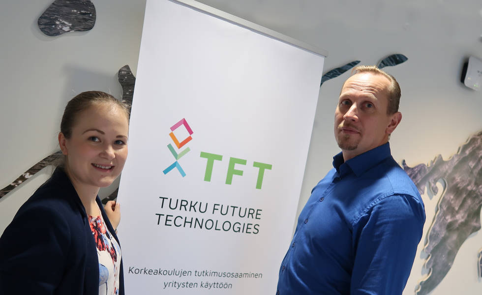 Sofia Tuominen ja Vesa Erkkilä seisovat TFT-tekstin ympärillä hymyillen.