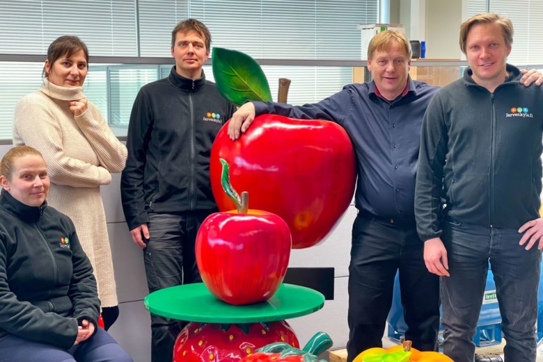 Viisi ihmistä seisoo kahden ison teko-omenan vieressä ja hymyilevät kameralle. 