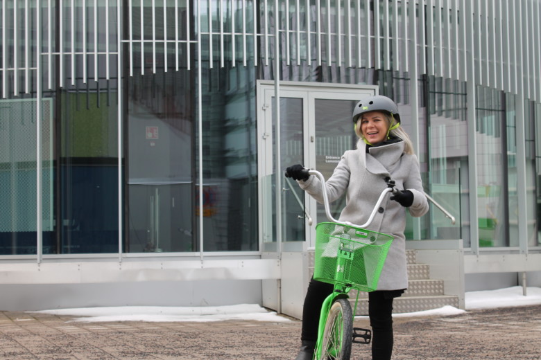 Noin 35-vuotias, pitkähiuksinen nainen hymyilee kameralle vihreän polkupyörän selässä. 