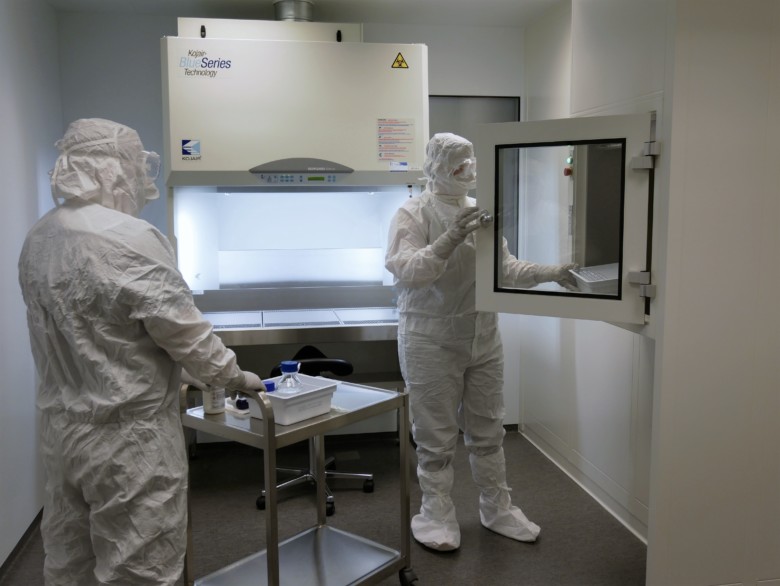 Two people dressed in white protective suits transfer manufacturing material in glass bottles from the hatch to the clean room. 