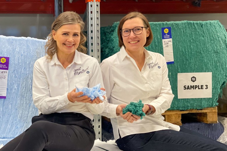 Two women, one wearing glasses, display blue and green recycled fibre in their hands. A green and blue fibre bale are visible behind them. 