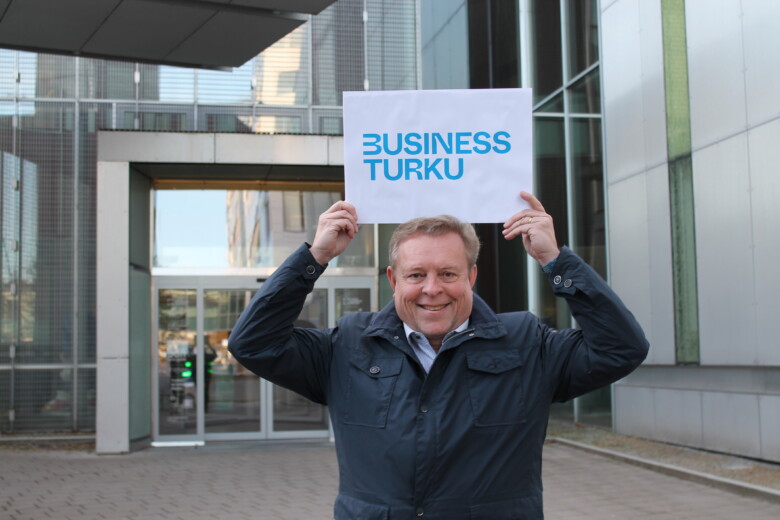 Kuvassa Business Turun toimitusjohtaja Tom Palenius pitää päänsä päällä kylttiä, jossa lukee sinisellä Business Turku. Takana on Joukahaisenkadulla sijaitsevan ICT-Cityn pääovi, josta pääsee Business Turun tiloihin. 